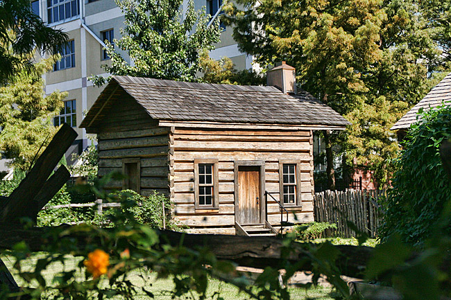 small log cabin in fenced-in plot with more modern city building, trees in background