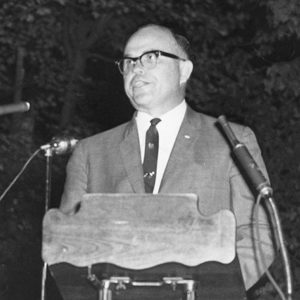 White man with glasses in suit and tie speaking at lectern