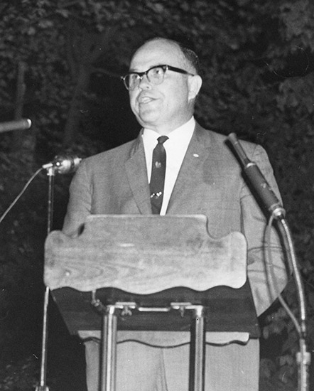 White man with glasses in suit and tie speaking at lectern