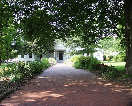 Wide brick sidewalk under tree-lined approach to white wood frame home