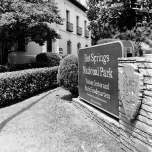 "Hot Springs National Park" sign with trees and two-story bath house with hospital building in the background