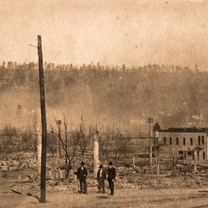 Three men standing in vacant lot with rubble next to storefronts and house