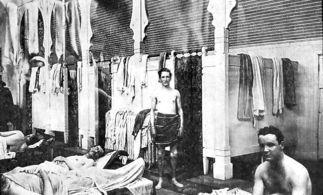Shirtless white men in bath house with stalls and beds