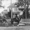White man in horse drawn carriage with African-American driver in front of multiple story house and fence
