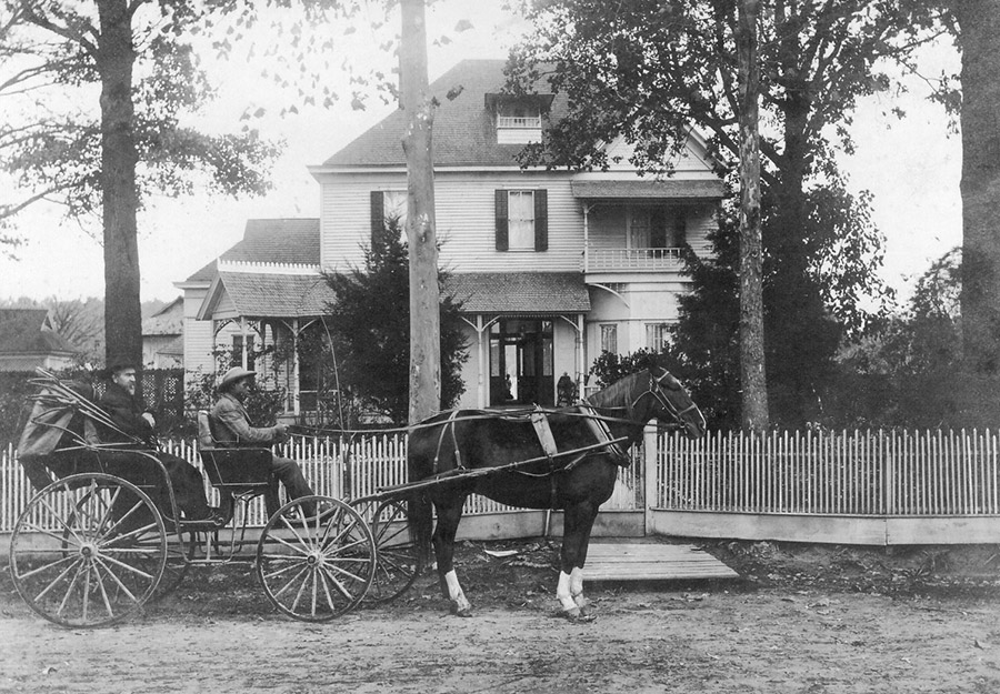 White man in horse drawn carriage with African-American driver in front of multiple story house and fence