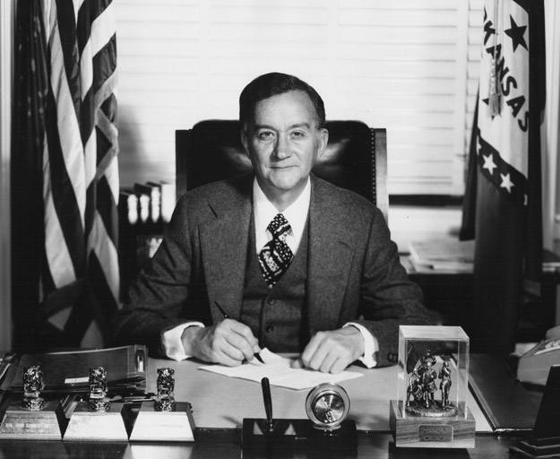Portrait white man in suit and tie smiling at desk signing paper flanked by U.S. and Arkansas flags