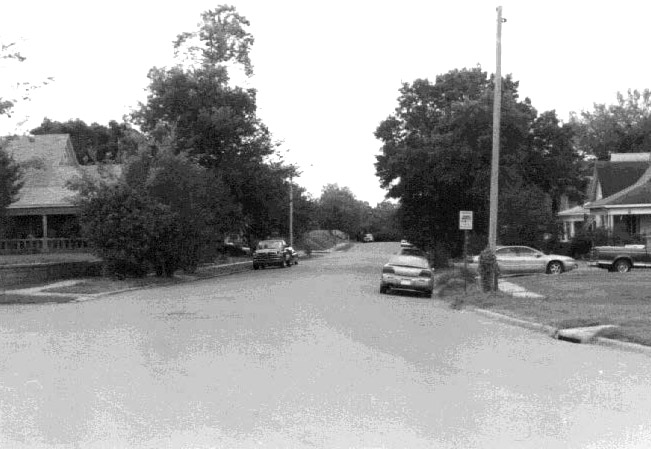 Looking down residential street with cars parked outside houses with covered porches and trees