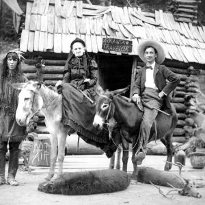 White man and old white woman on donkeys with white woman on foot dressed in Native American garb in front of log cabin with "Arkansaw Traveler" sign above the door