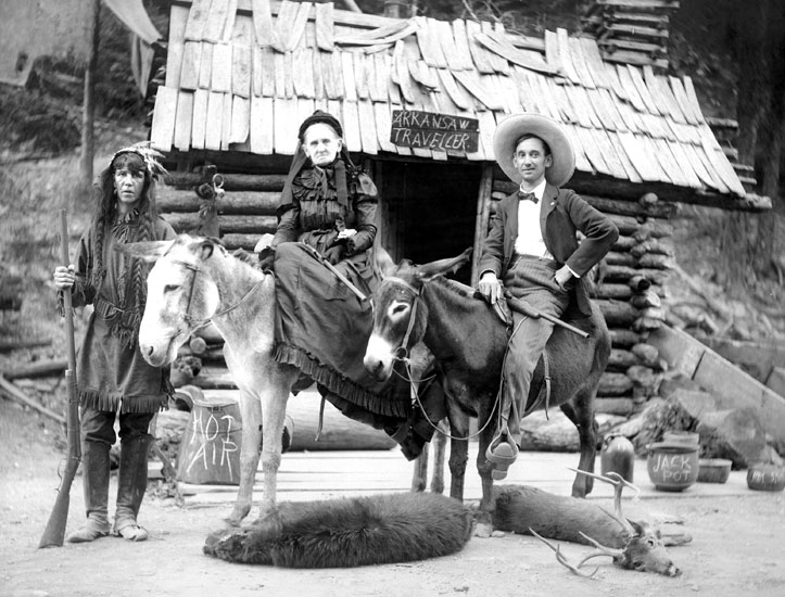 White man and old white woman on donkeys with white woman on foot dressed in Native American garb in front of log cabin with "Arkansaw Traveler" sign above the door