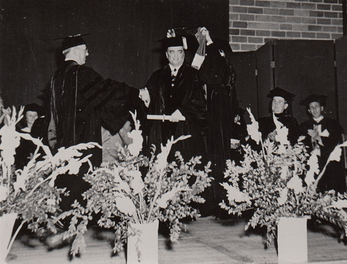 Graduation ceremony with young white man being hooded while shaking hands with older white man in robes on stage
