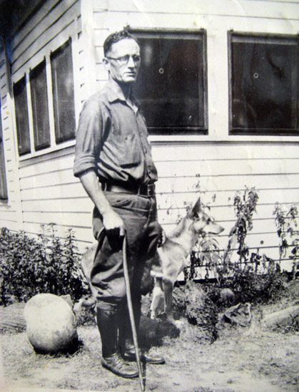 White man with cane and dog standing outside at corner of a house