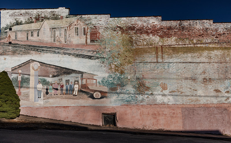 Faded mural depicting train depot building and service station with patrons and car on brick building