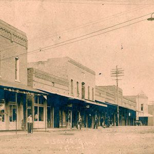 Men standing under awnings outside multistory brick storefront buildings on town street