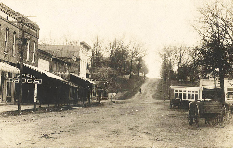 Looking down dirt street with single and multistory storefront buildings and wagons in the foreground