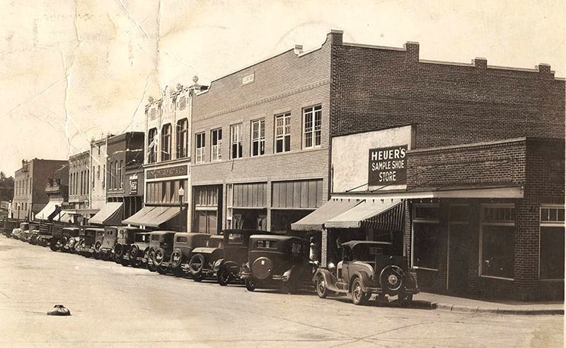 Row of cars parked on street outside multistory and single-story storefront buildings