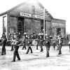 Group of white men in uniforms with instruments marching past brick storefronts in street