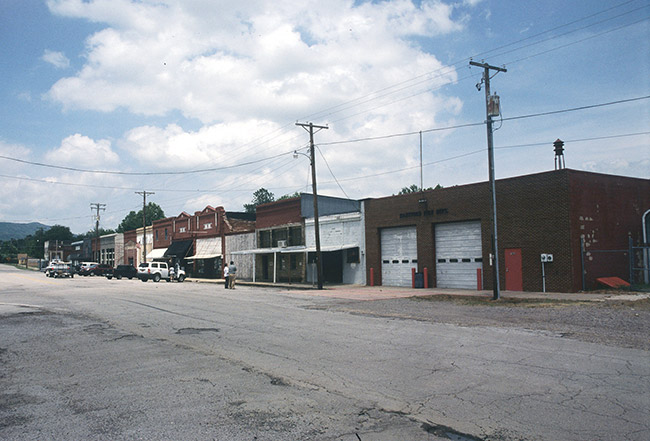 Street with brick storefronts and two-bay brick garage building with parked cars and two people standing