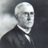 Older white man with round glasses in suit and tie