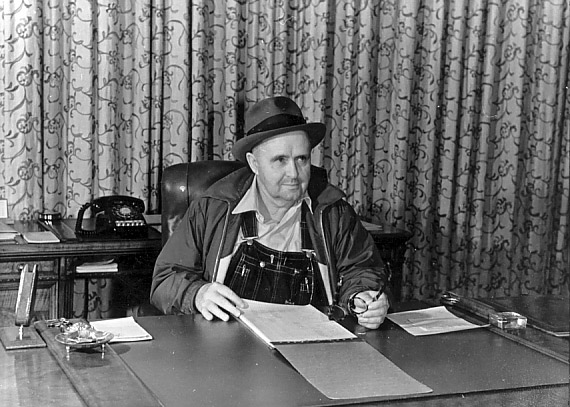 white man in overalls wind breaker and fedora sitting at desk with curtains behind him
