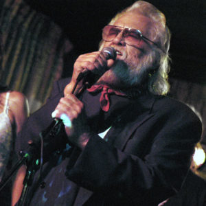 Older white man with beard and glasses singing at microphone with younger white woman on stage