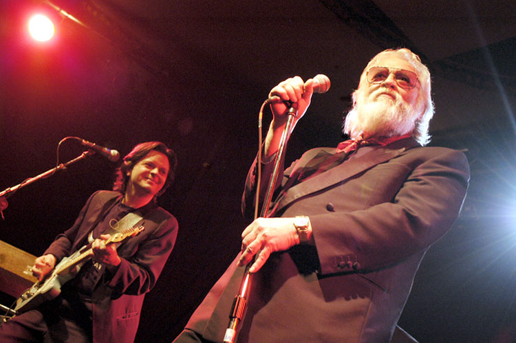 Older white man with beard at microphone on stage with younger white guitarist