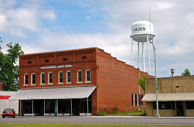 Two-story brick store building with awning and single-story storefront on street with water tower in the background