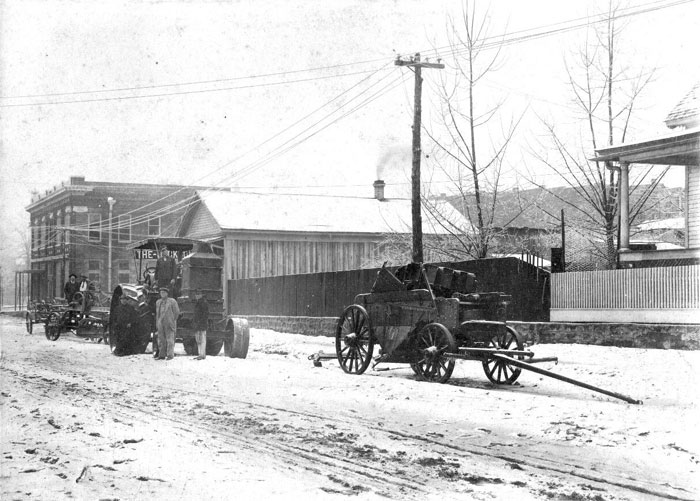 White men driving a tractor on a dirt road with buildings and telephone poles in the background