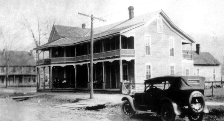 Car parked on street near two-story building featuring wraparound porches on both levels with telephone poles and another two-story building in the background