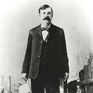 White man with bushy mustache standing with hat in hand at wooden fence