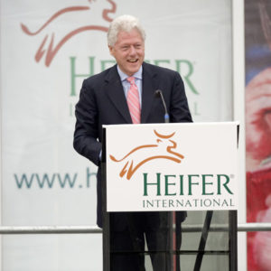 President Bill Clinton in suit tie smiling at "Heifer International" lectern on stage banner background