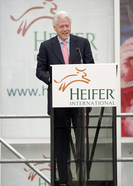 President Bill Clinton in suit tie smiling at "Heifer International" lectern on stage banner background