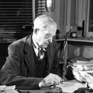 Old white man in suit and tie writing at a cluttered desk