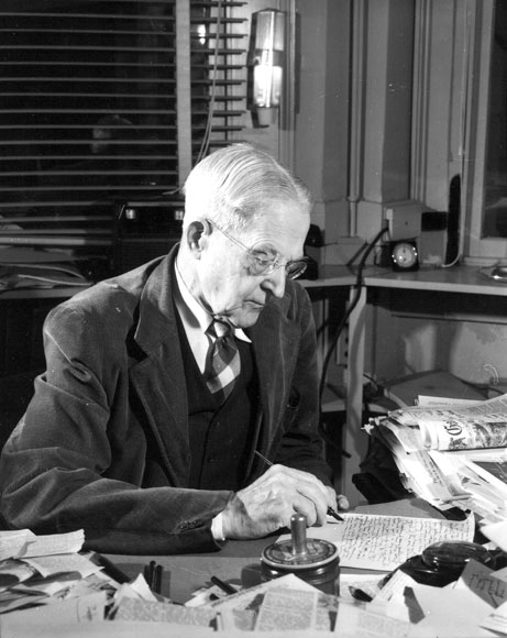 Old white man in suit and tie writing at a cluttered desk