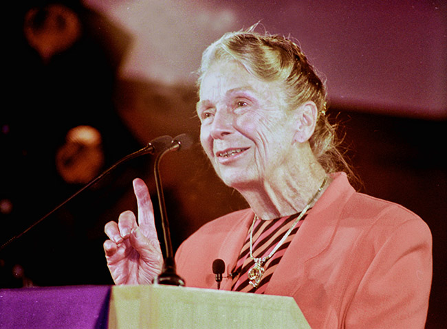 Older white woman with pulled back hair in a pink and black striped top and pink jacket talking at a lectern