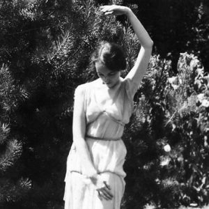 Young white woman dancing by a tree in white dress with her left arm extended above her head