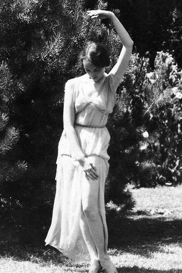 Young white woman dancing by a tree in white dress with her left arm extended above her head