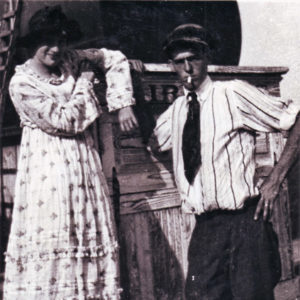 White man in shirt and tie smoking a cigarette with white woman in dress and hat