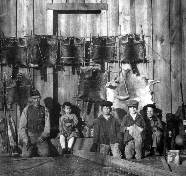 White man and children posing with raccoon hides hanging stretched on wooden walls