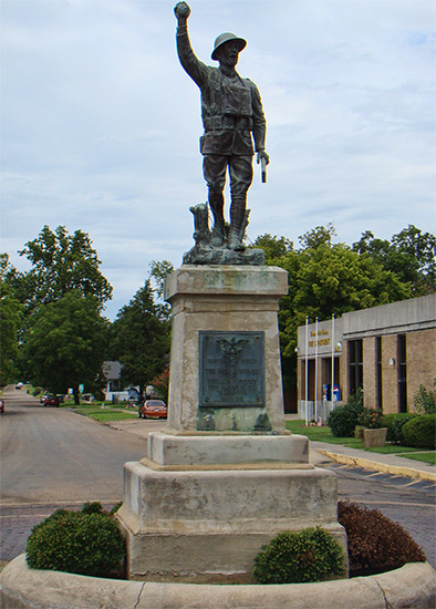 Statue of soldier with his left hand raised on stone pedestal with plaque