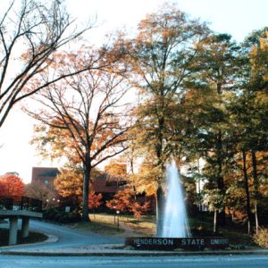 "Henderson State University" sign by fountain by campus entrance driveway with tall trees brick and cement architecture