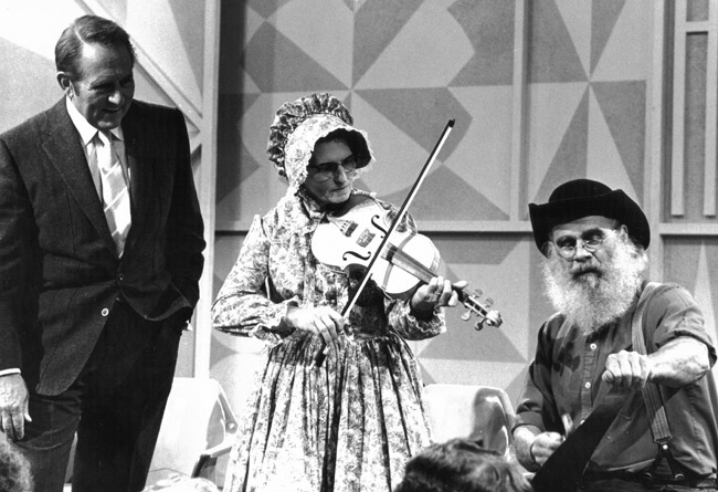 White man in suit smiling observing white woman and man playing violin and musical saw