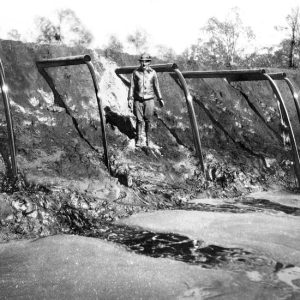 Old white man standing next to a dirt wall from which protrude pipes pouring oil into a pit
