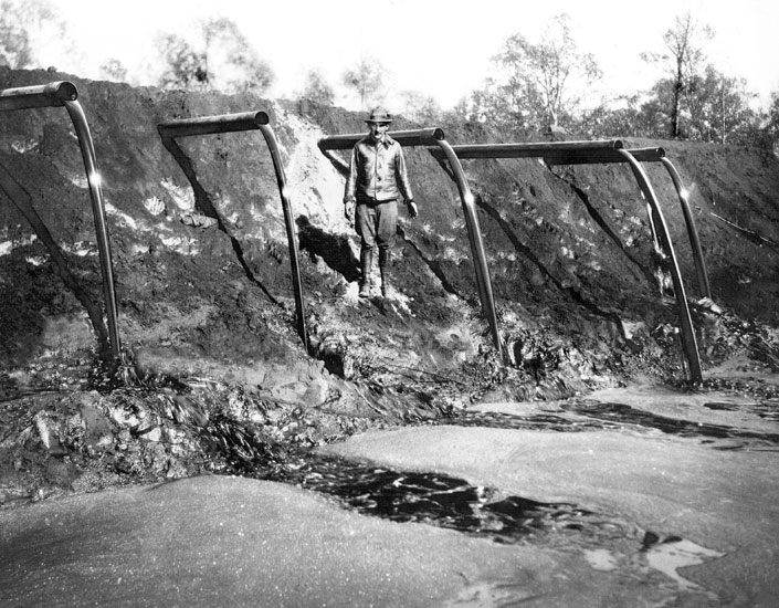 Old white man standing next to a dirt wall from which protrude pipes pouring oil into a pit