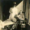 White man in wheelchair smoking a cigarette with white nurse standing over him holding his wrist