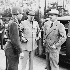 White man in hat and suit standing by car with open door talking to white men in military uniforms
