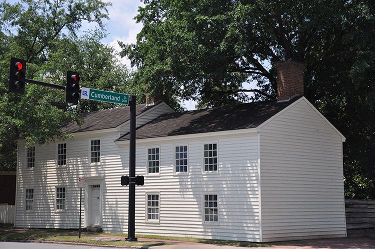 Two-story building with white siding and brick chimney on street corner next to "Cumberland" traffic light