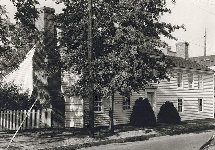 Two-story building with white siding and brick chimney behind tree on street corner