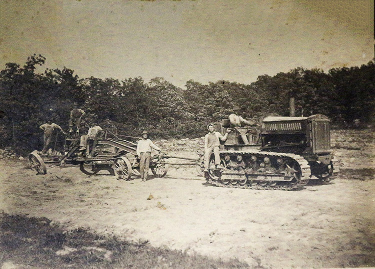 faded photo of men pulling machinery with tractor with trees in the background