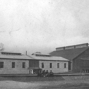 Group of white men sitting outside long single-story building with barn-like structure beside it
