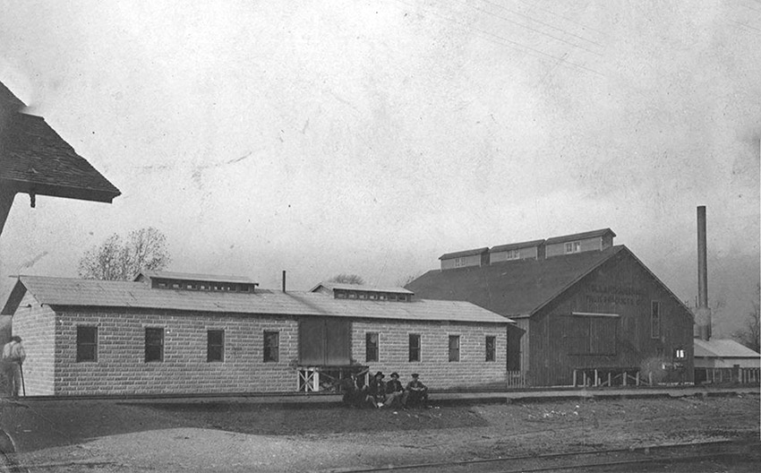 Group of white men sitting outside long single-story building with barn-like structure beside it